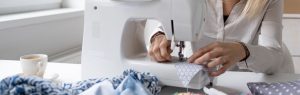 A woman is using a sewing machine. There is blue material on the table in front of the machine. Slow fashion, or making your own clothes, is good for your mental health and wellbeing, as well as the planet in terms of sustainability and reducing carbon footprint.